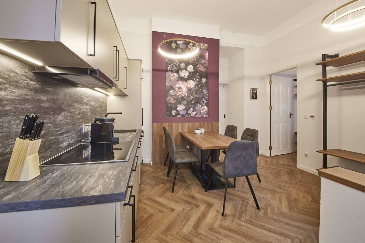A modern kitchen with a wooden dining table and chairs, a hanging light fixture, and a herringbone-patterned floor.