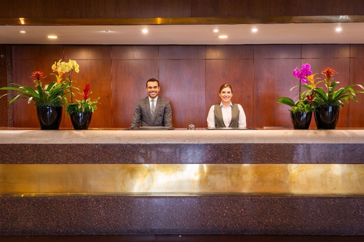A reception desk with two individuals seated, surrounded by potted plants.