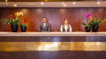 A reception desk with two individuals seated, surrounded by potted plants.