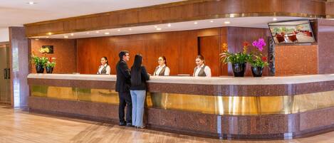 A hotel reception desk with a marble counter, wooden paneling, and potted plants.