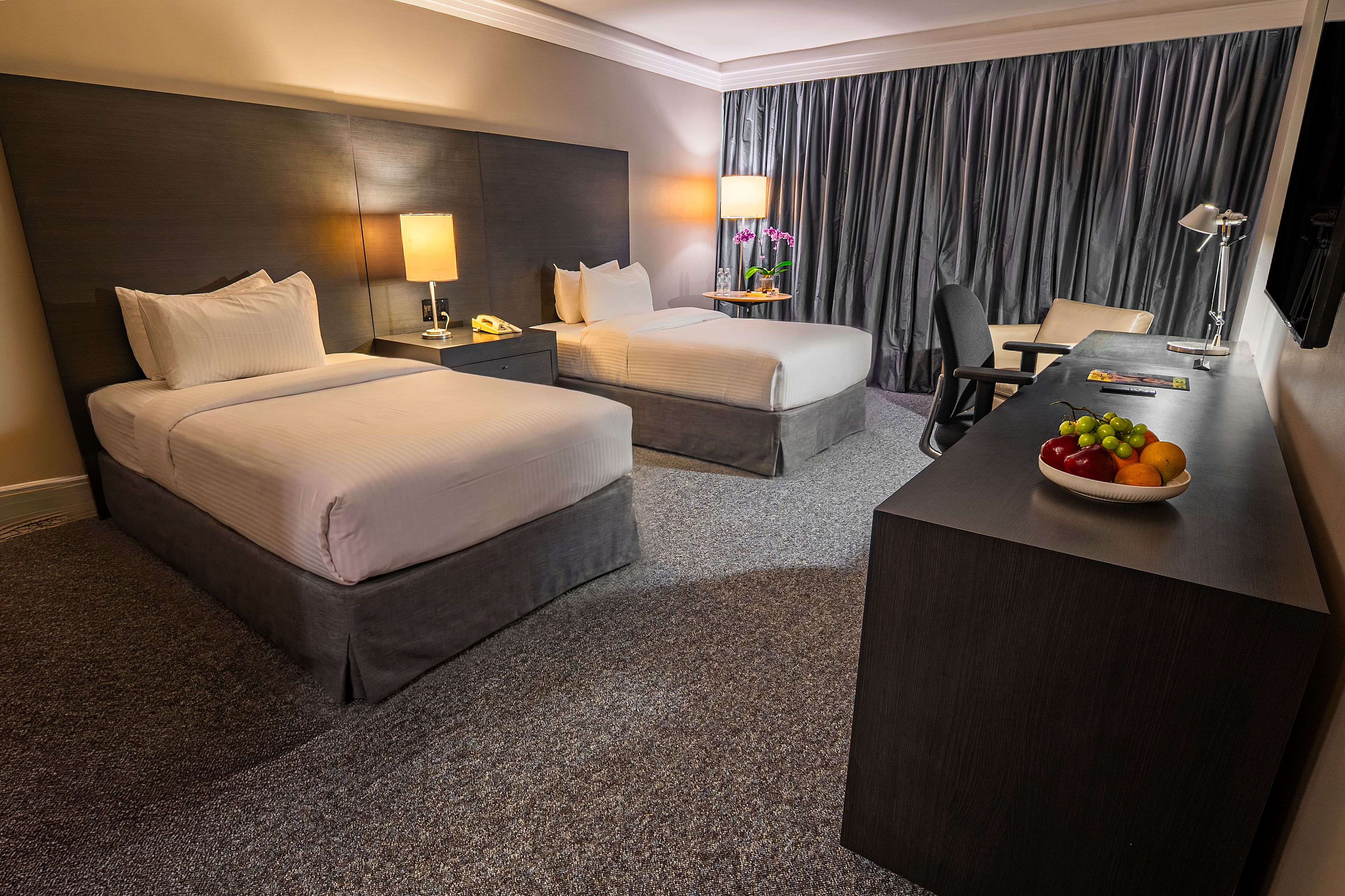 A hotel room with two beds, a desk with a fruit bowl, a lamp, and a view of the outside.