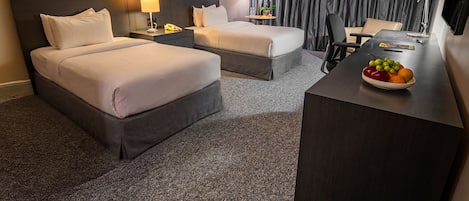 A hotel room with two beds, a desk with a fruit bowl, a lamp, and a view of the outside.