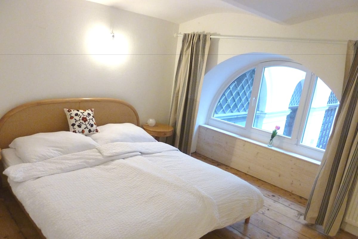 A bedroom with a large bed, wooden floor, a nightstand, a lamp, a window with curtains, and a view of buildings outside.