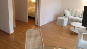 Une chambre d’hôtel moderne avec un plancher en bois, une table en verre décorée de bougies, un fauteuil en osier, un canapé à rayures et un lit avec une tête de lit blanche.