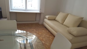 Sala de estar com um sofá bege, uma mesa de vidro e uma janela com vista para edifícios.