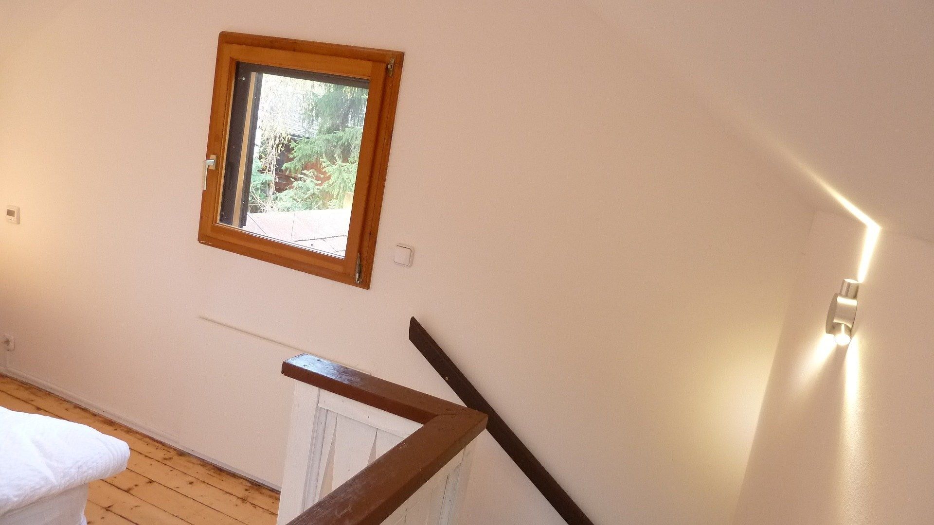 Ein Zimmer mit Holzboden, einem weißen Heizkörper, einer Holztreppe, einem kleinen Fenster mit Blick auf Grünflächen und einer Wandleuchte.