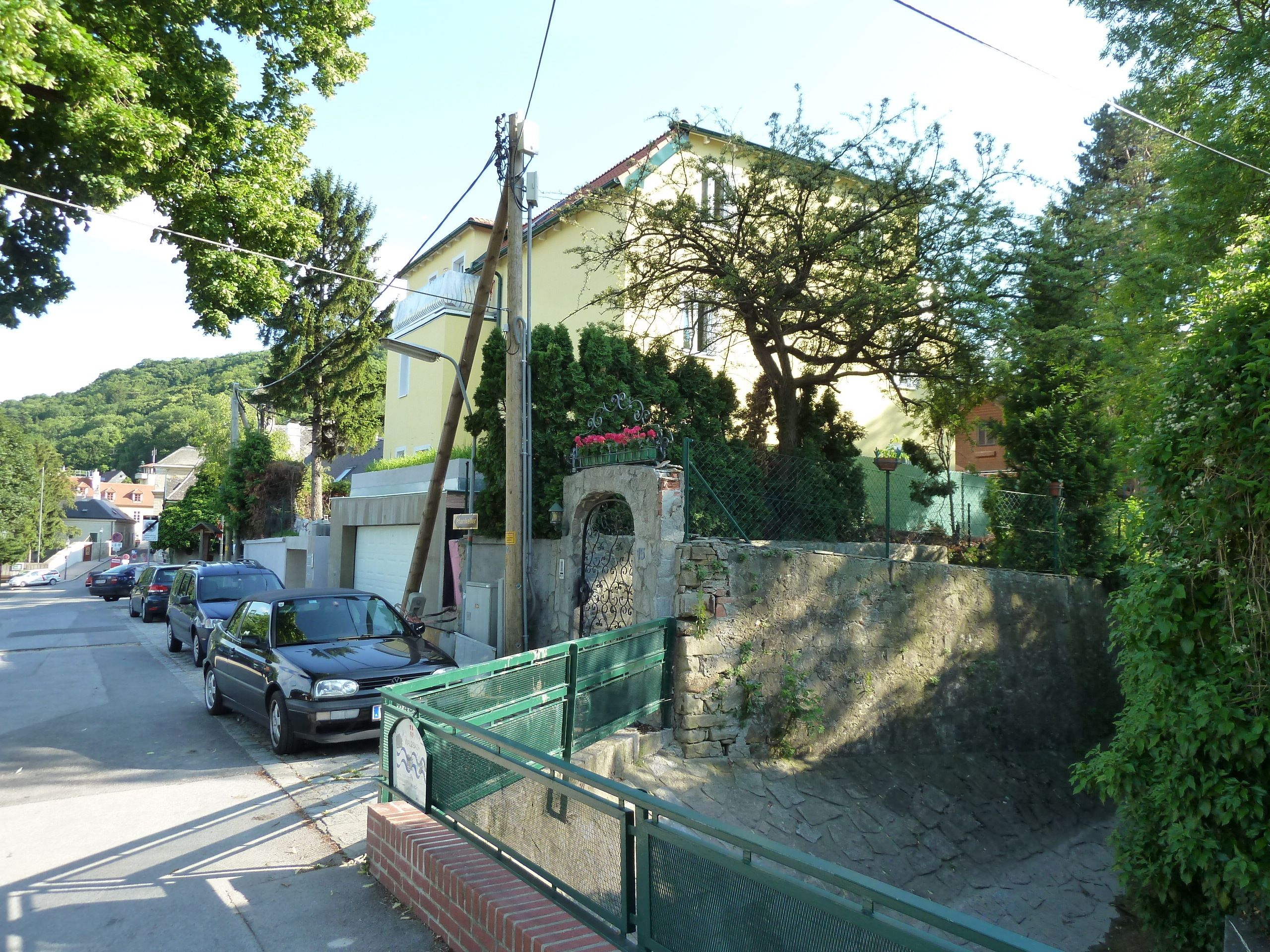 A residential street with parked cars, a stone wall, and a green metal fence.