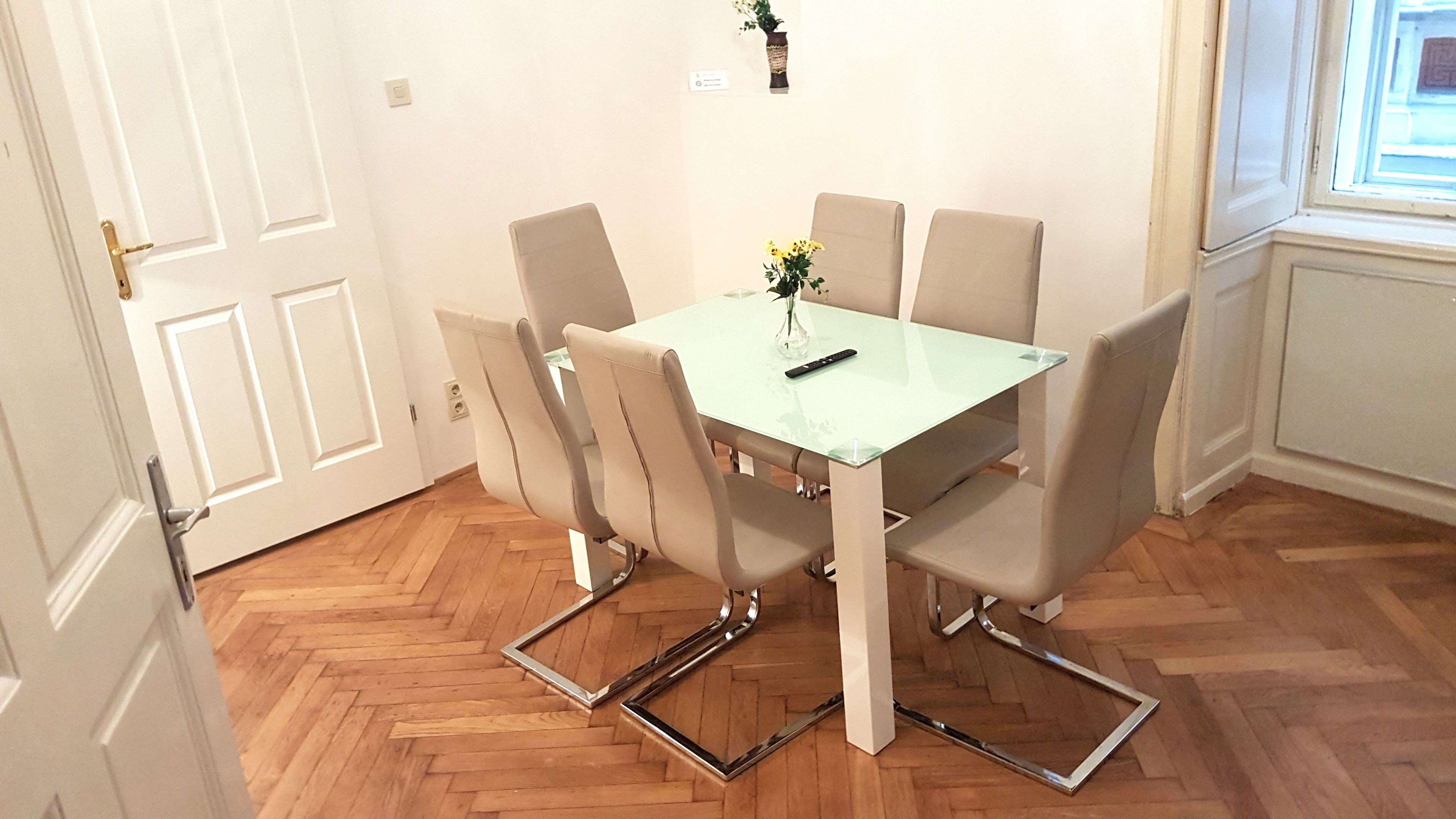 A dining area with a glass table and four chairs, a vase with flowers, and a remote control on the table.