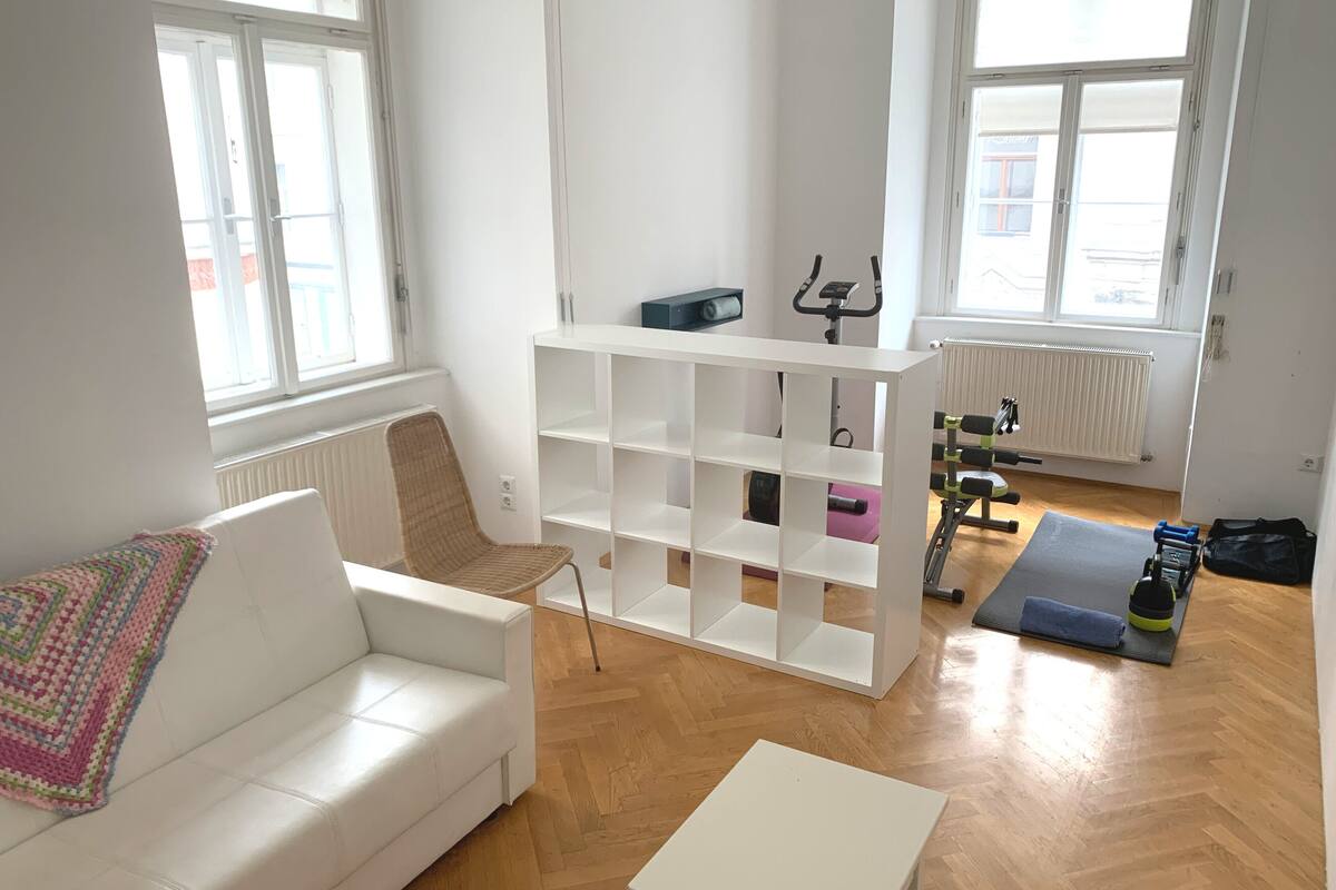 Una sala de estar con un sofá blanco, una mesita pequeña, una silla de mimbre, una estantería blanca y una colchoneta de yoga con equipo de ejercicios.