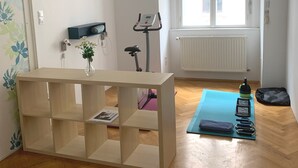Una habitación con piso de madera, una bicicleta estática, una colchoneta de yoga y una pequeña mesa con un florero y una revista.