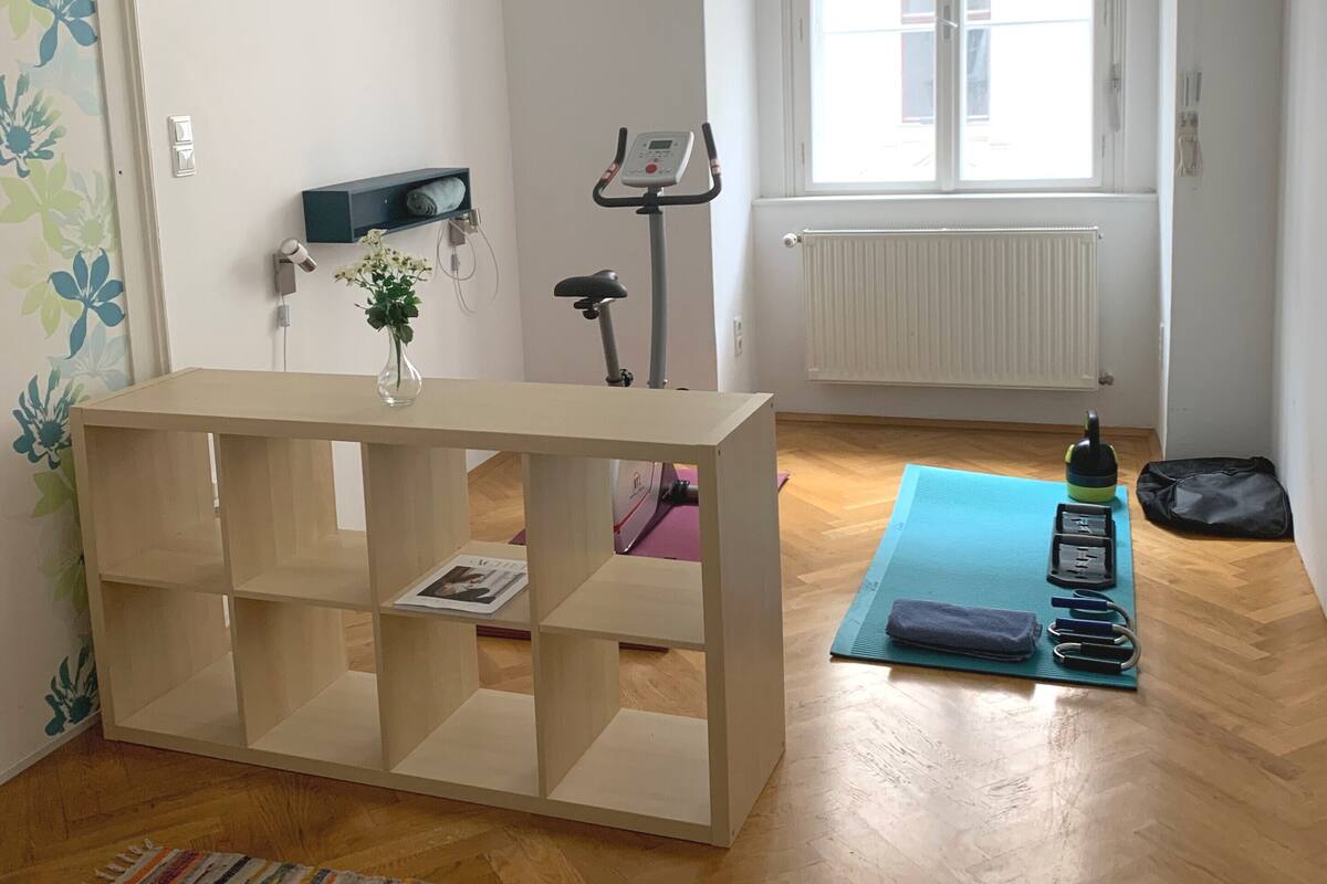 Una habitación con piso de madera, una bicicleta estática, una colchoneta de yoga y una pequeña mesa con un florero y una revista.