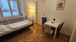 Una habitación pequeña y ordenada con una cama individual, una mesa con una silla, una lámpara y un cuadro en la pared.