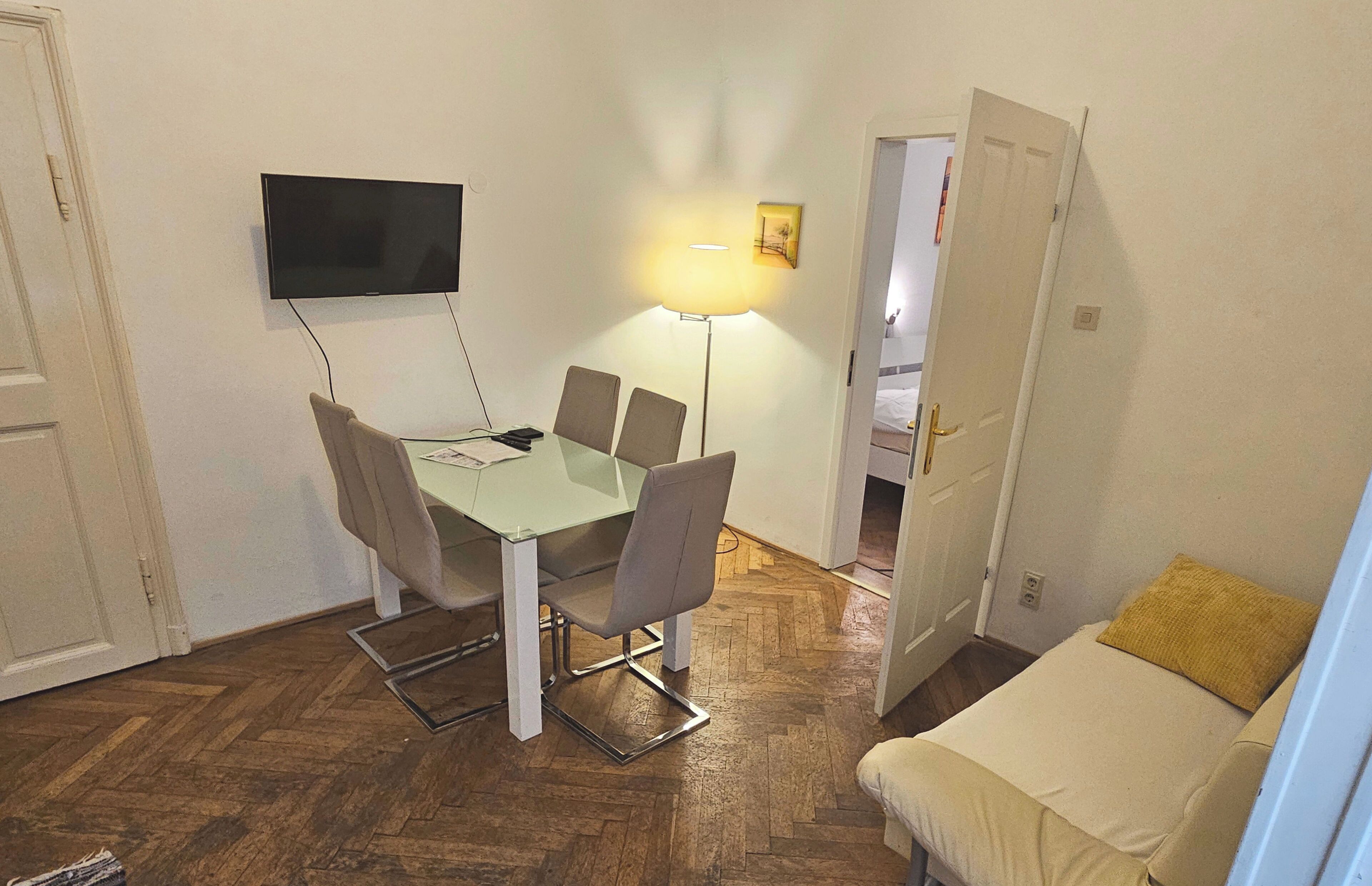 A compact room with a dining table, chairs, a TV mounted on the wall, a floor lamp, a bed with a pillow, and a door leading to another room.