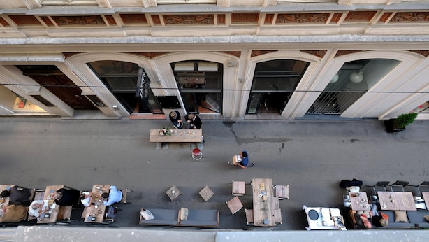 Un café al aire libre con mesas y sillas dispuestas frente a un edificio con ventanas amplias y arcos decorativos.