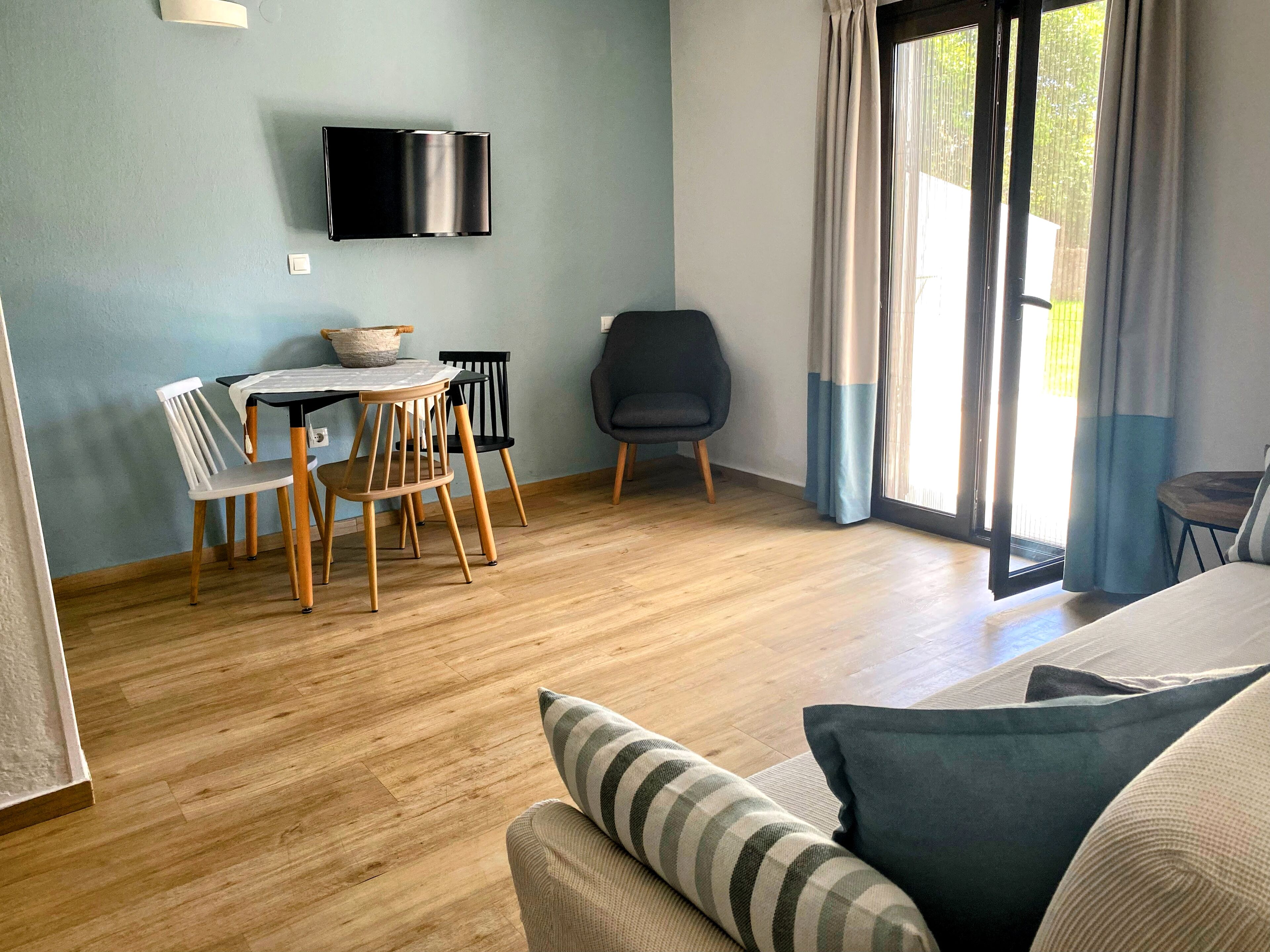 A modern room with a wooden floor, a dining table with chairs, a black chair, a sofa with cushions, a flat-screen TV mounted on the wall, and a sliding glass door with curtains.