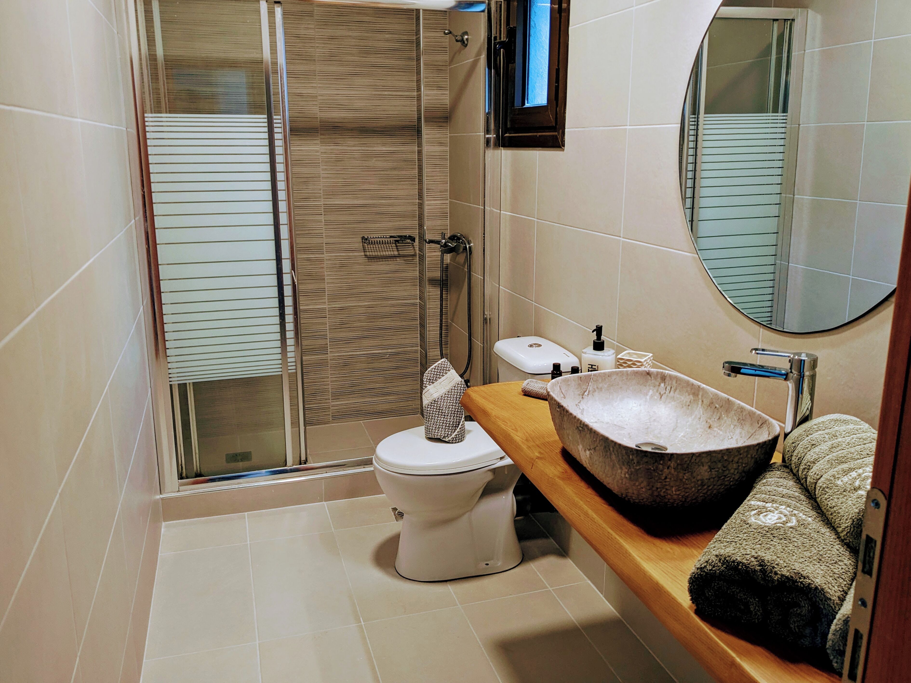 A modern bathroom with a glass shower enclosure, a white toilet, a wooden vanity with a large basin sink, and a round mirror.