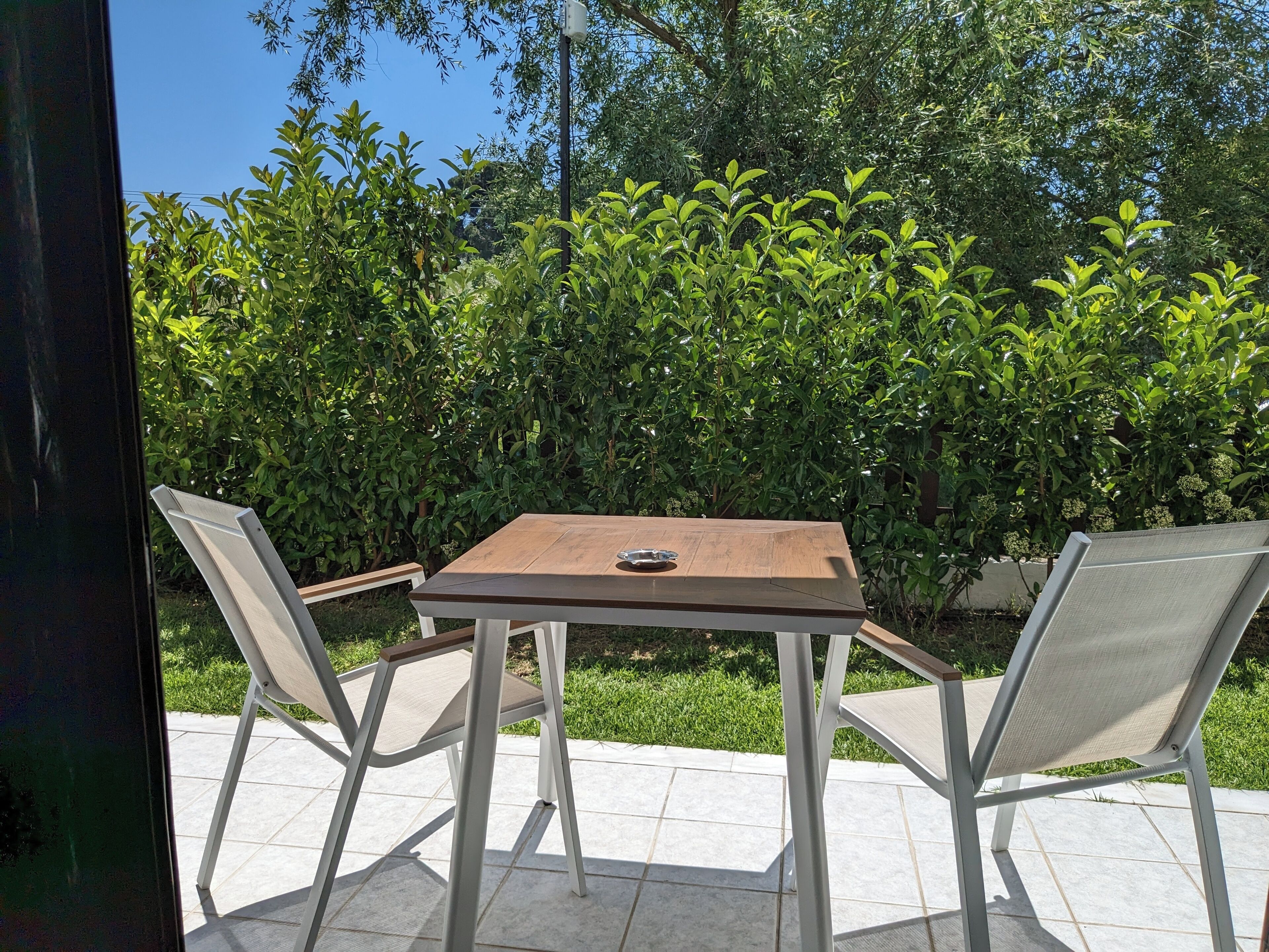 An outdoor patio with a wooden table and two chairs, surrounded by lush greenery.