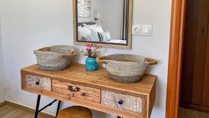A wooden console table with two woven baskets and a vase with flowers, above a mirror reflecting a room with a bed and an air conditioner.