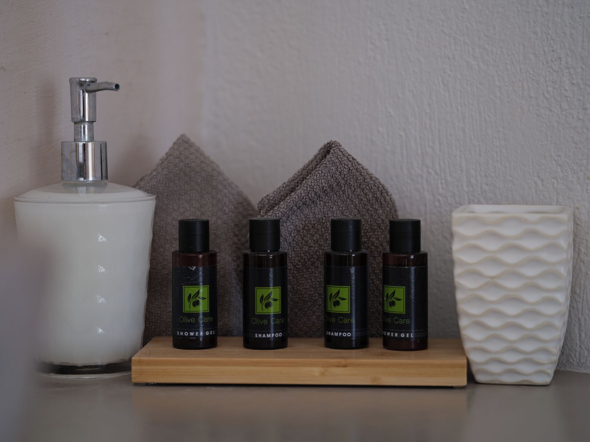 A bathroom shelf with a showerhead, four bottles of Olive Care products, and a textured white vase.