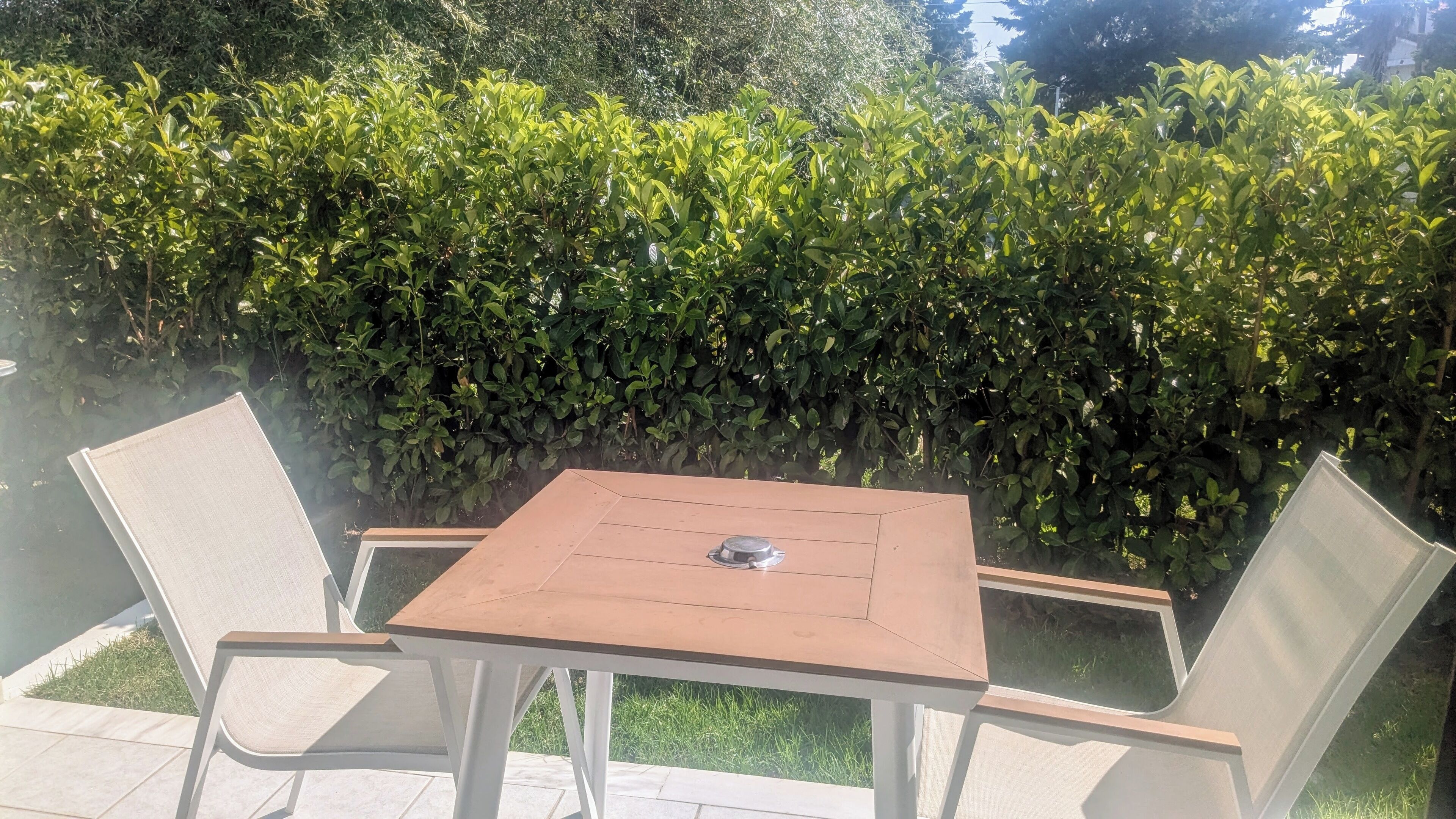 An outdoor patio with a square table and two chairs, surrounded by a hedge.