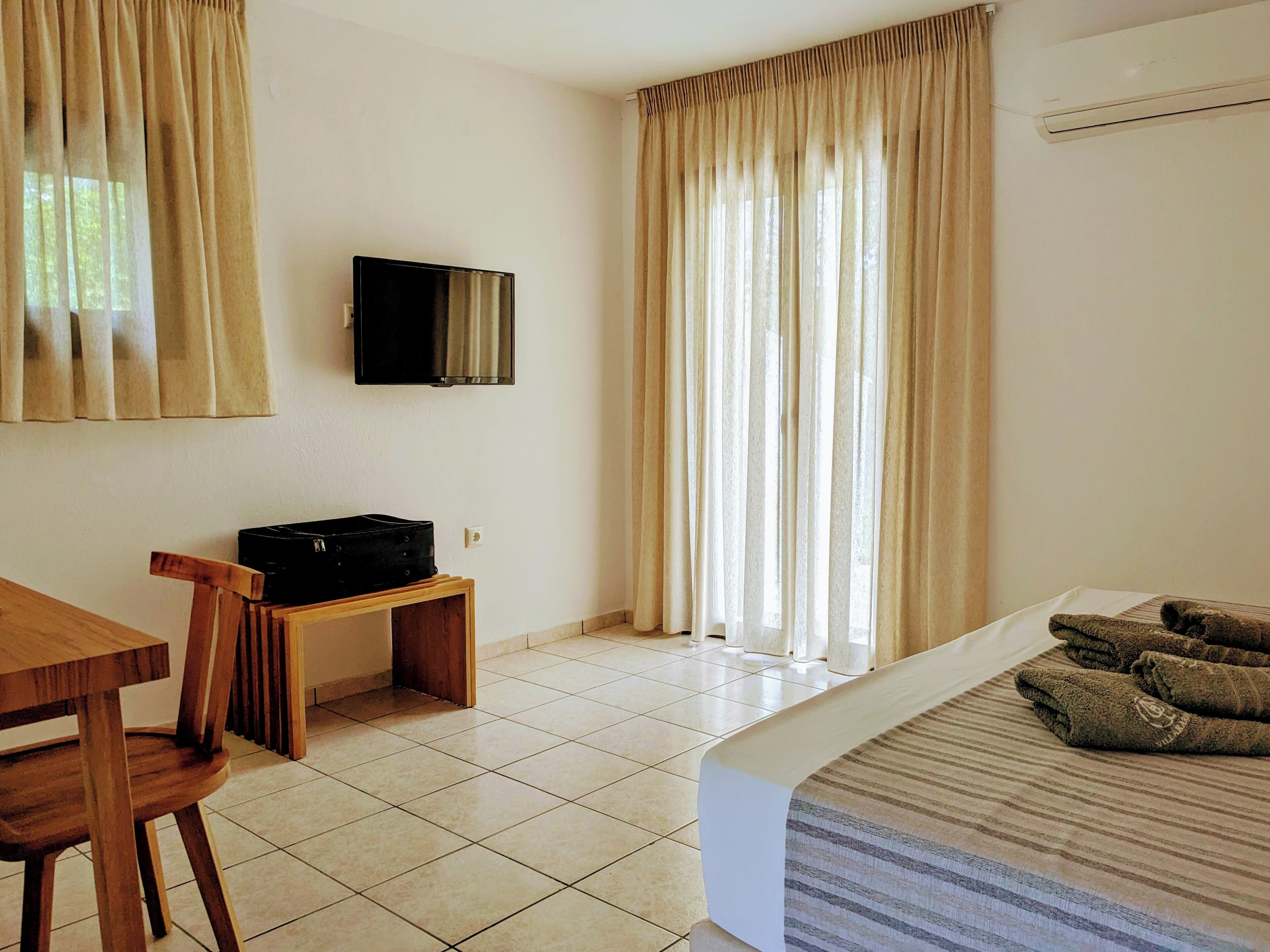 A hotel room with a bed, a TV mounted on the wall, a desk with a chair, and a window with curtains.