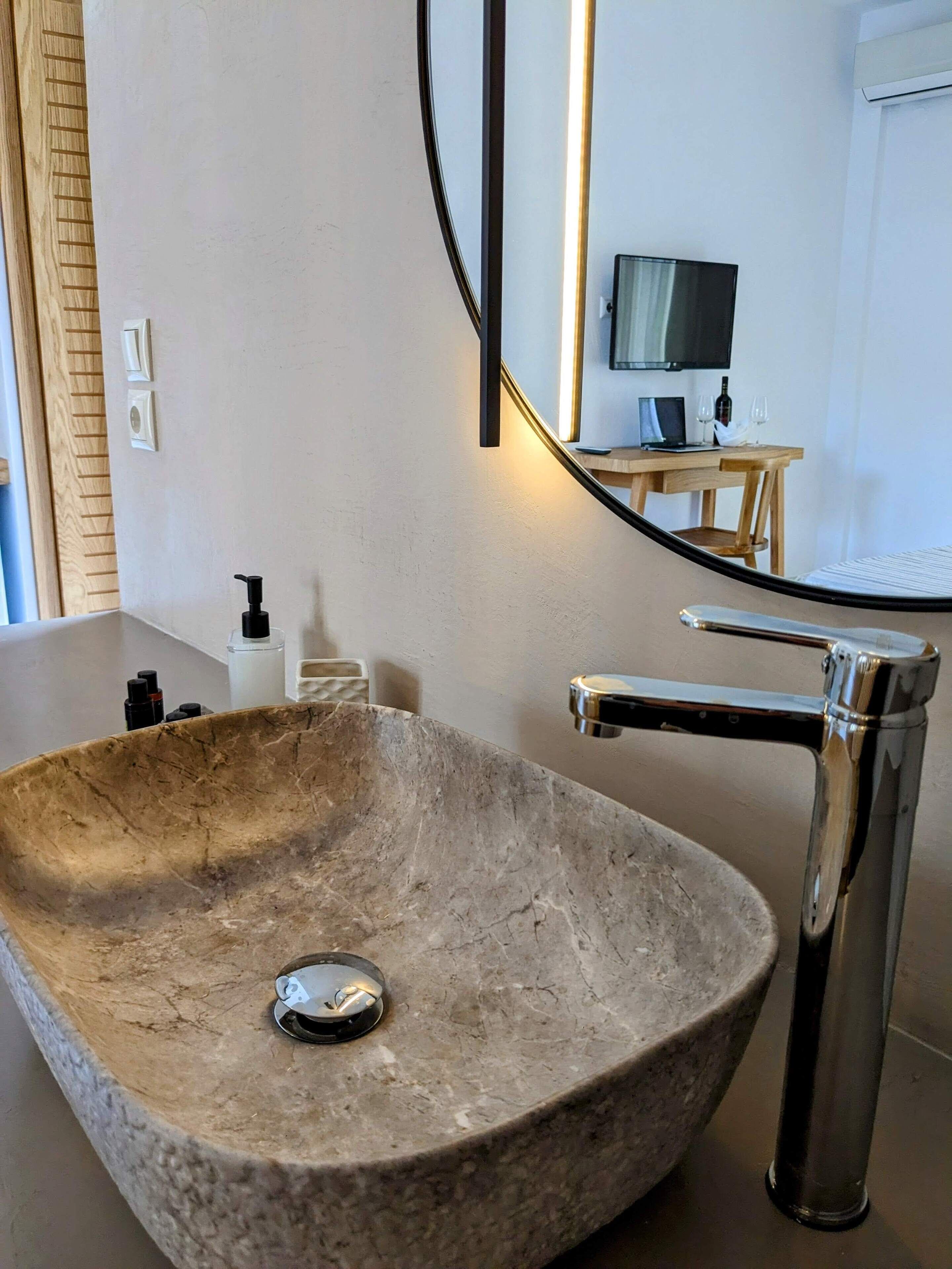 Une salle de bain moderne avec un grand lavabo en pierre, un robinet chromé et un miroir qui reflète un téléviseur et une petite table avec des verres à vin.