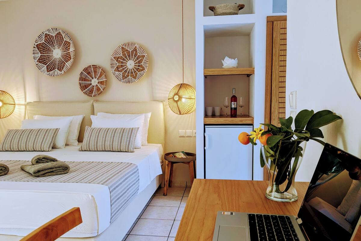 Ein Schlafzimmer mit einem Bett, einem Nachttisch und einem Schreibtisch mit einem Laptop. Auf dem Schreibtisch befindet sich ein kleiner Kühlschrank und ein Blumentopf.