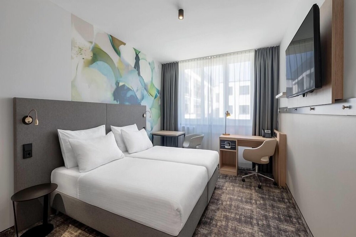 A hotel room with a bed, desk, chair, TV, and abstract wall art.