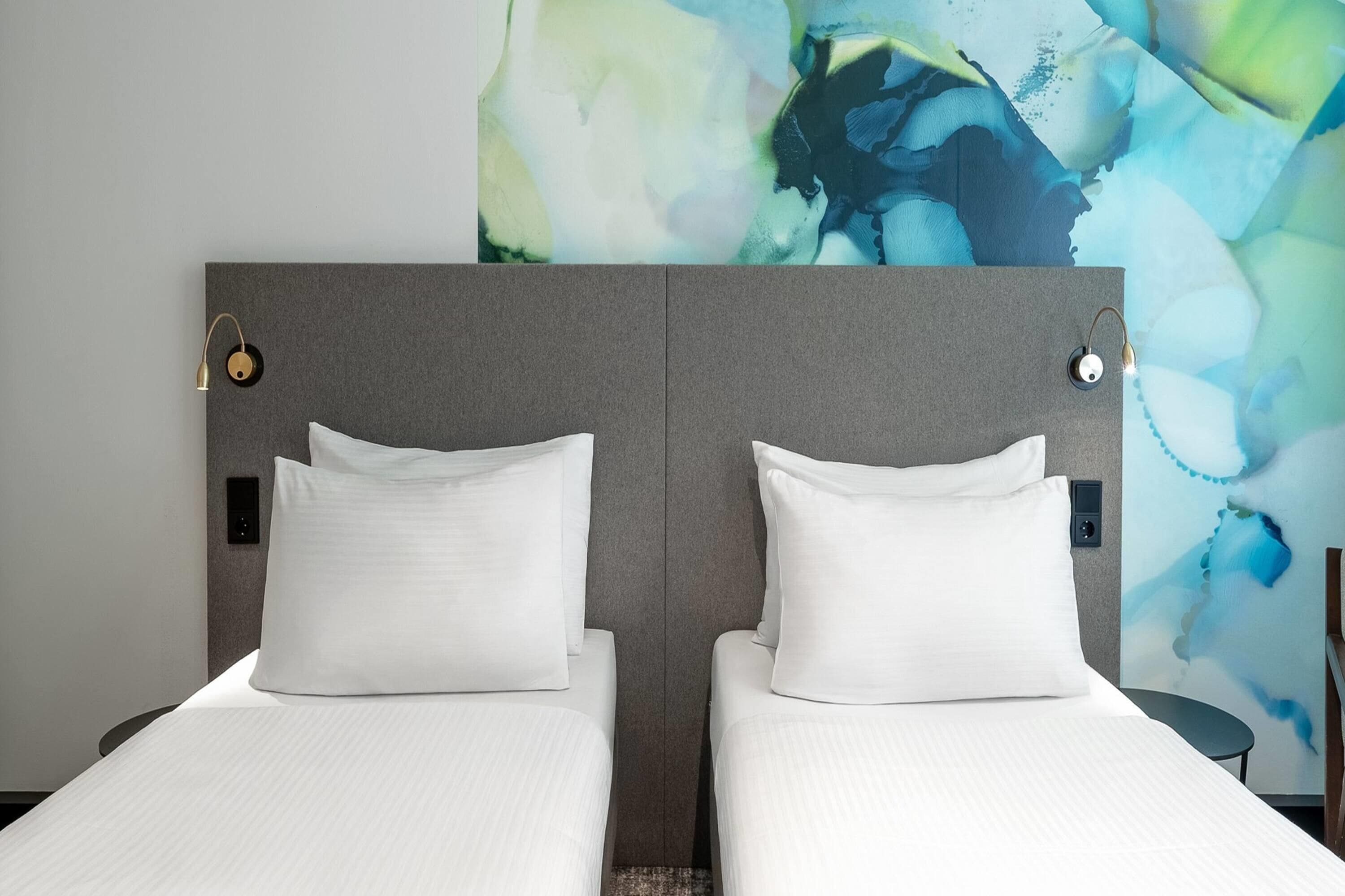 A double bed with white bedding and a grey headboard, a wall-mounted light, and a large abstract painting.