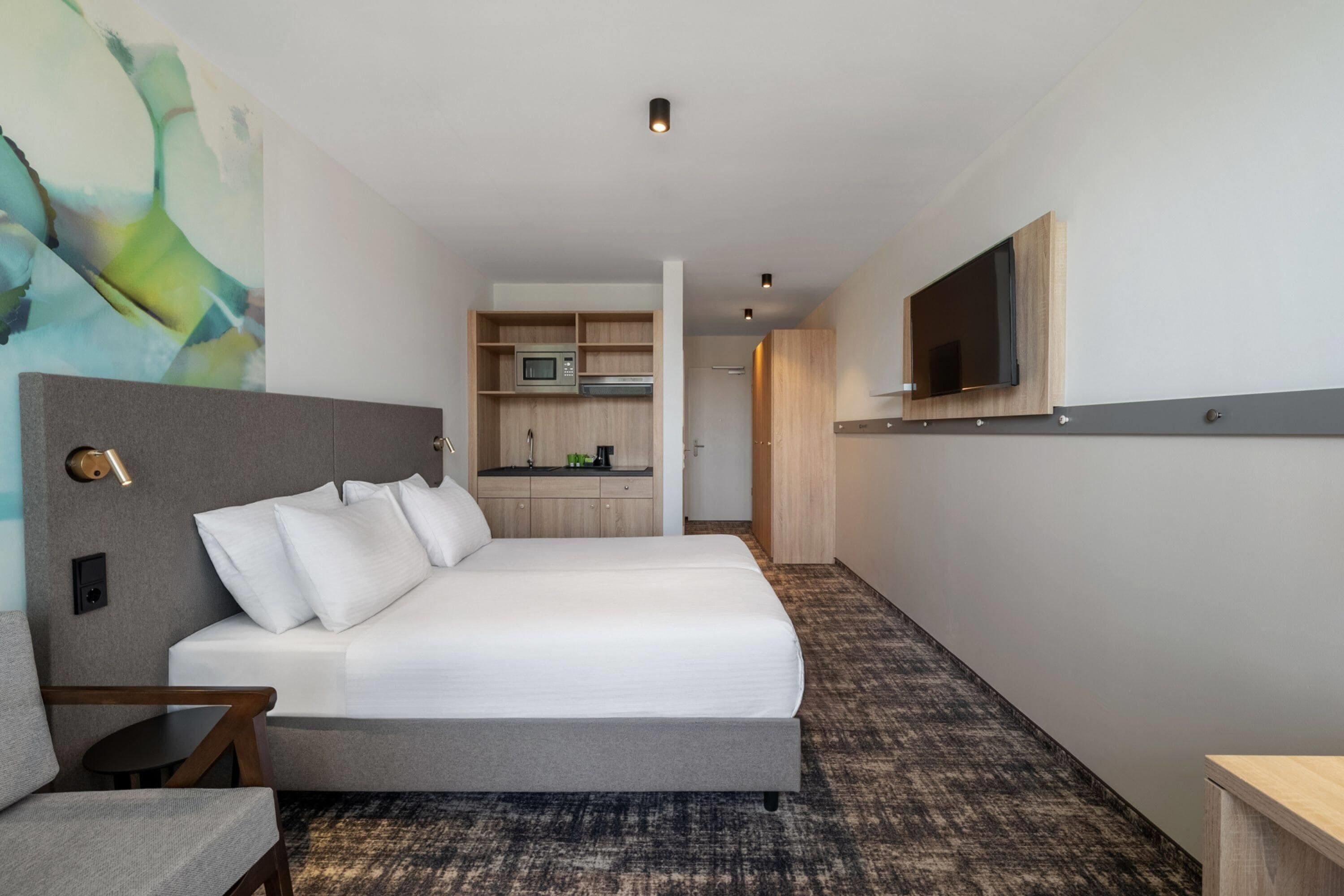 A hotel room with a bed, a small table, a chair, a TV mounted on the wall, and a kitchenette in the background.