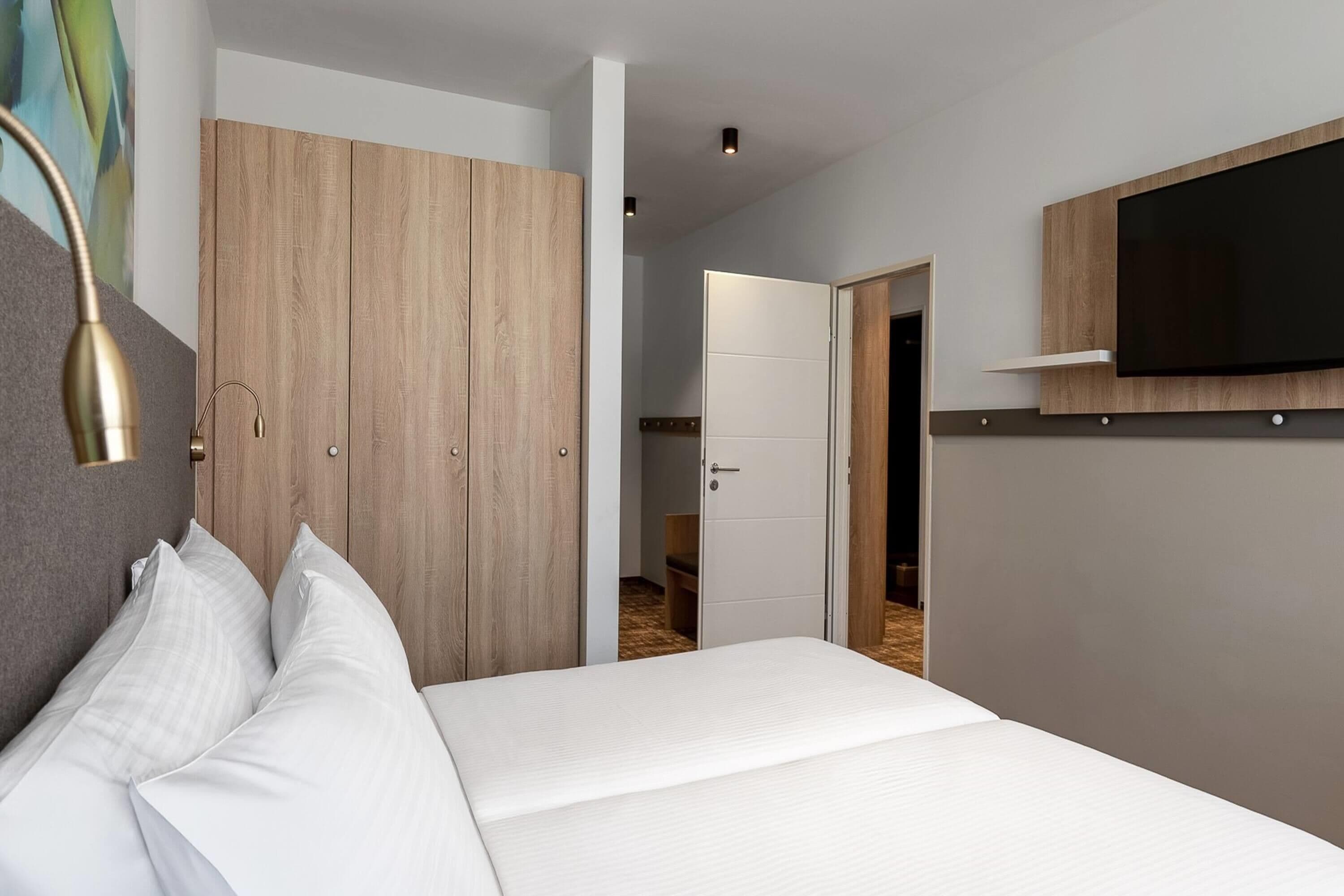 A modern bedroom with a bed, a wardrobe, a TV, and a door leading to a bathroom.