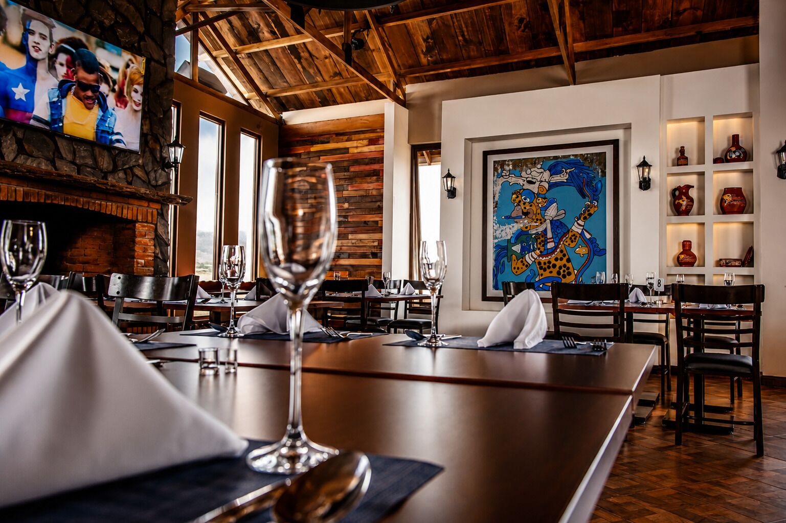 A restaurant interior with wooden beams, a large painting, and a television on the wall.