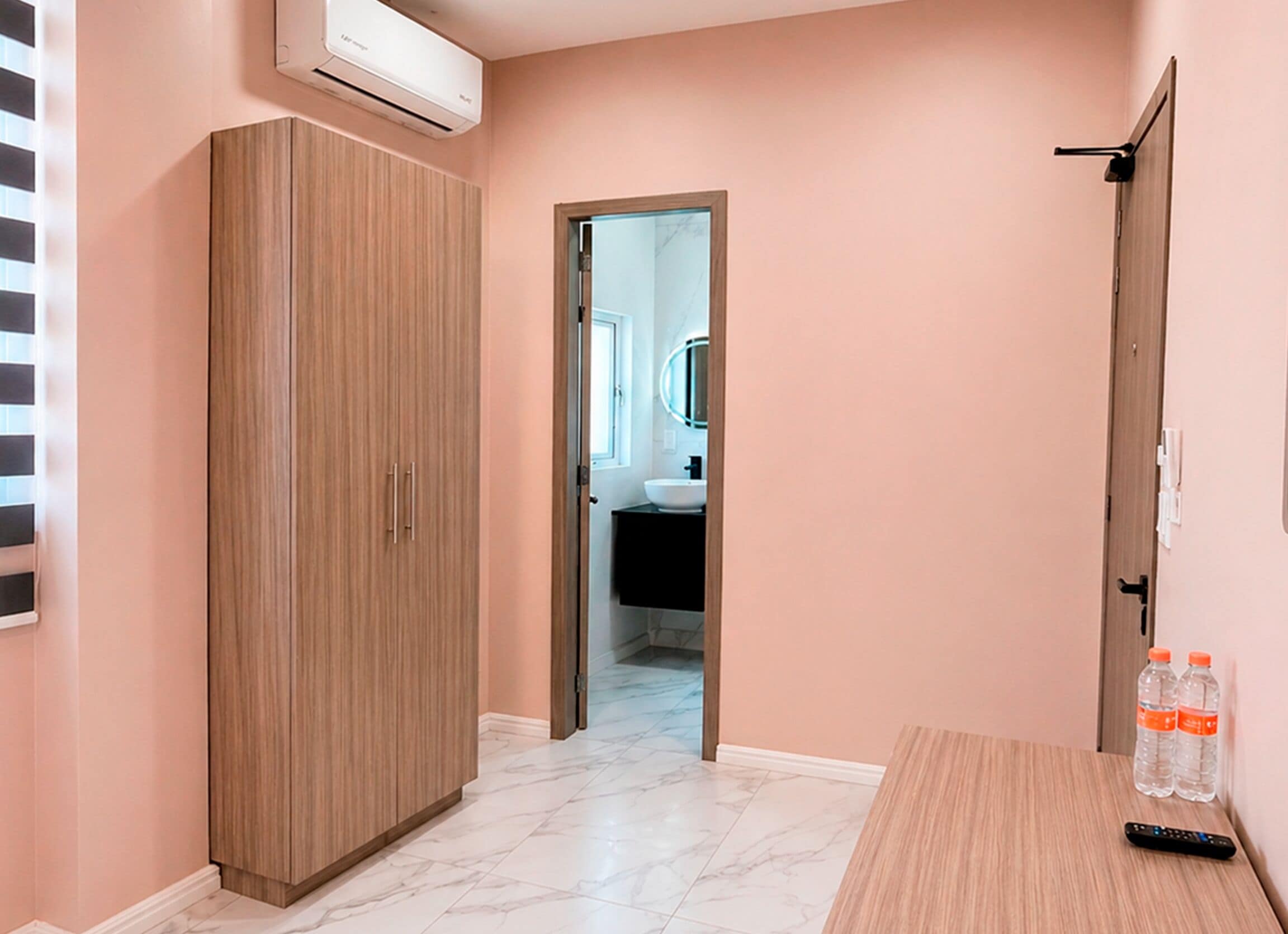 A hallway with a wooden wardrobe, a bathroom visible through an open door, and a table with two bottles and a remote control.