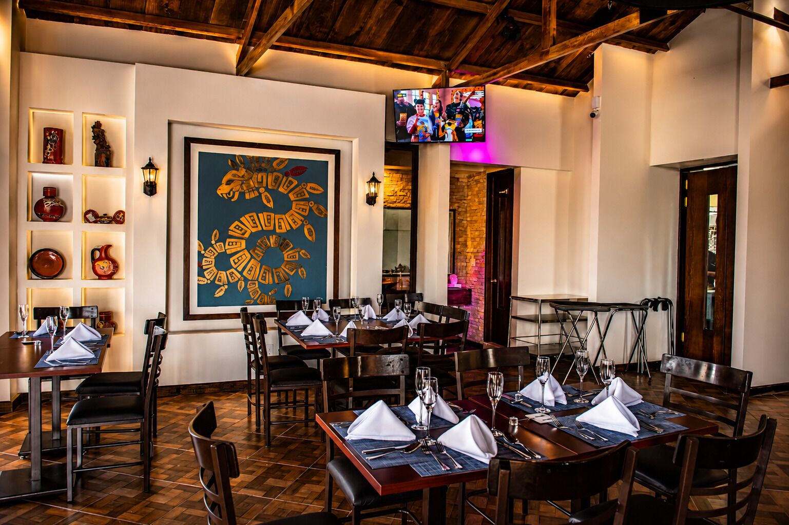 A restaurant with multiple tables set for dining, a large framed artwork on the wall, and a television screen above the tables.