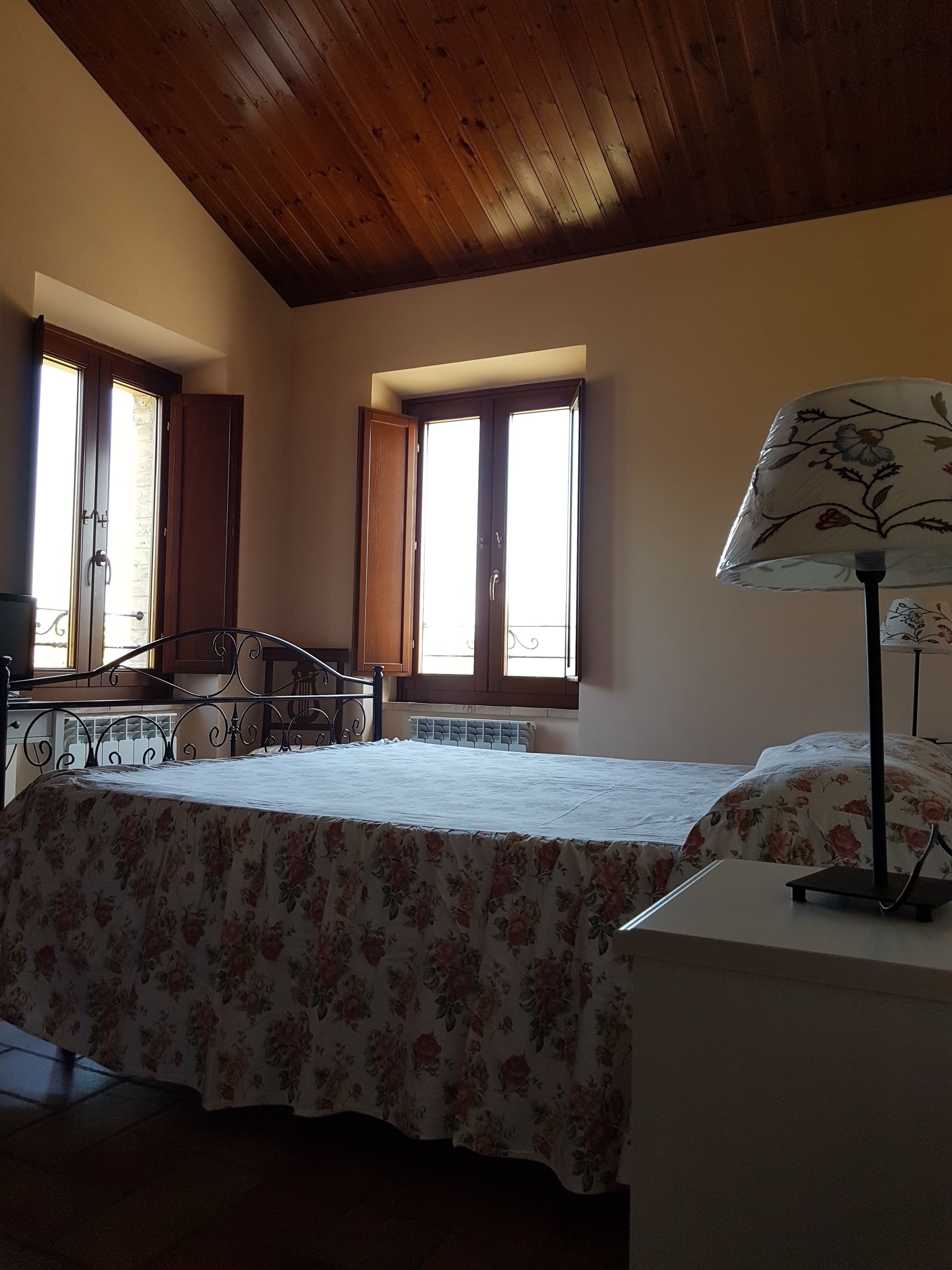 A bedroom with a wooden ceiling, a bed with a floral bedspread, a bedside table with a lamp, and two windows with curtains.