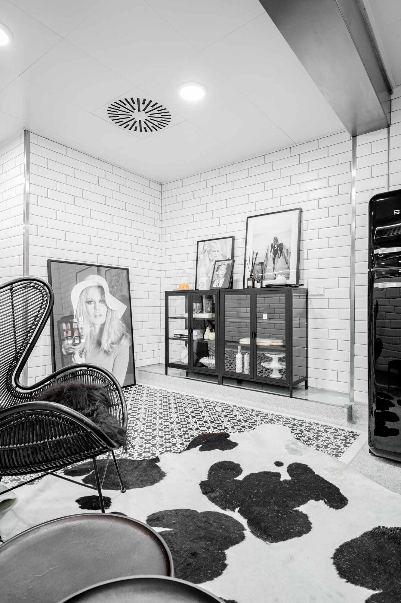 Una habitación moderna con paredes de ladrillo blanco, un tapete de cuero de vaca blanco y negro, una silla con estructura metálica, un aparador de vidrio con fotografías enmarcadas y una lámpara de techo.