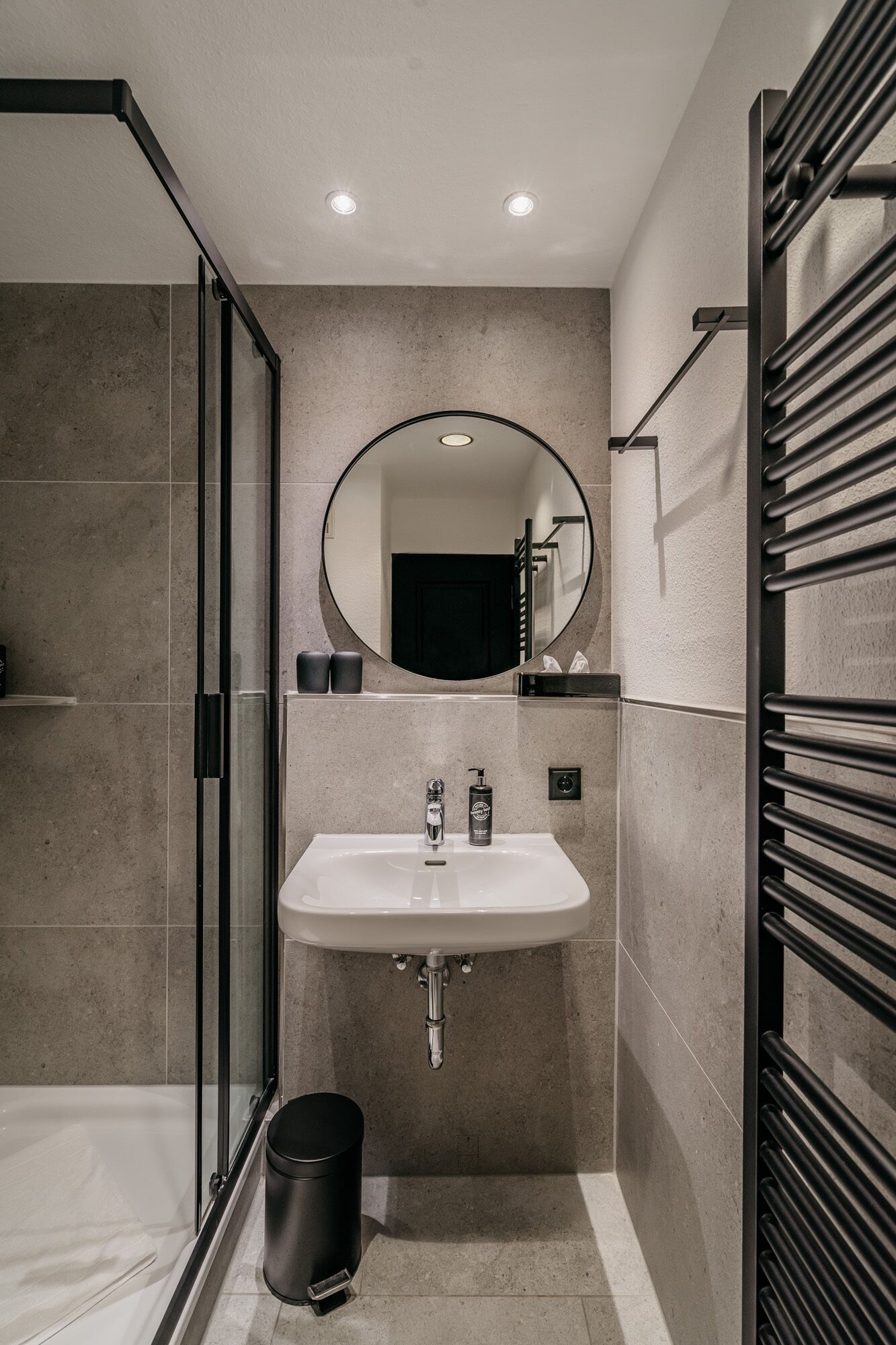 A modern bathroom with a glass shower enclosure, a white sink with a mirror above it, and a black trash bin.