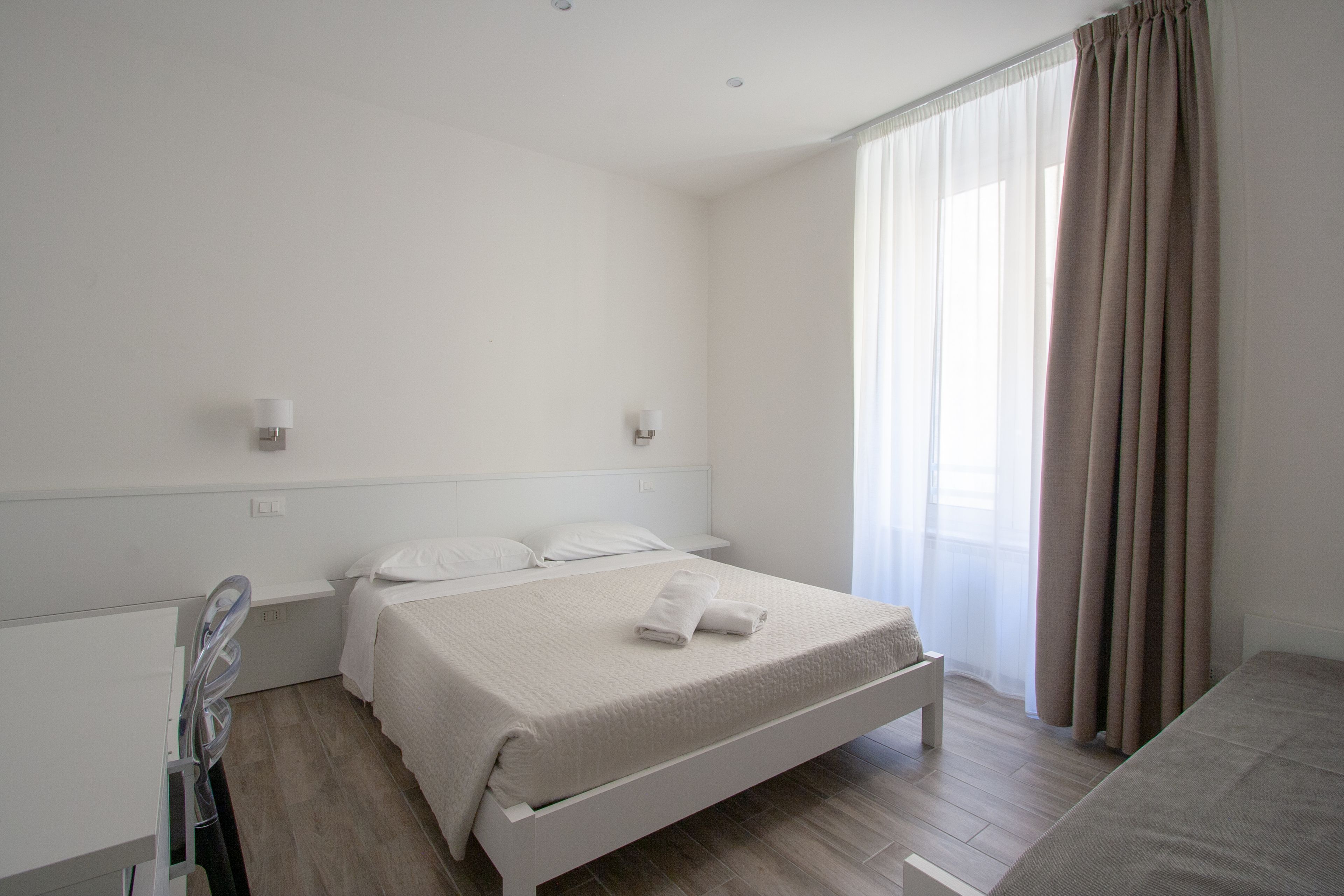 Quarto moderno e limpo, com uma cama de solteiro, mesa de cabeceira e uma janela grande com cortinas.