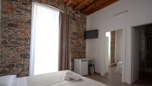 Ein Schlafzimmer mit einer Steinwand, einem Bett mit weißer Decke, einem Schreibtisch und einem an der Wand befestigten Fernseher.