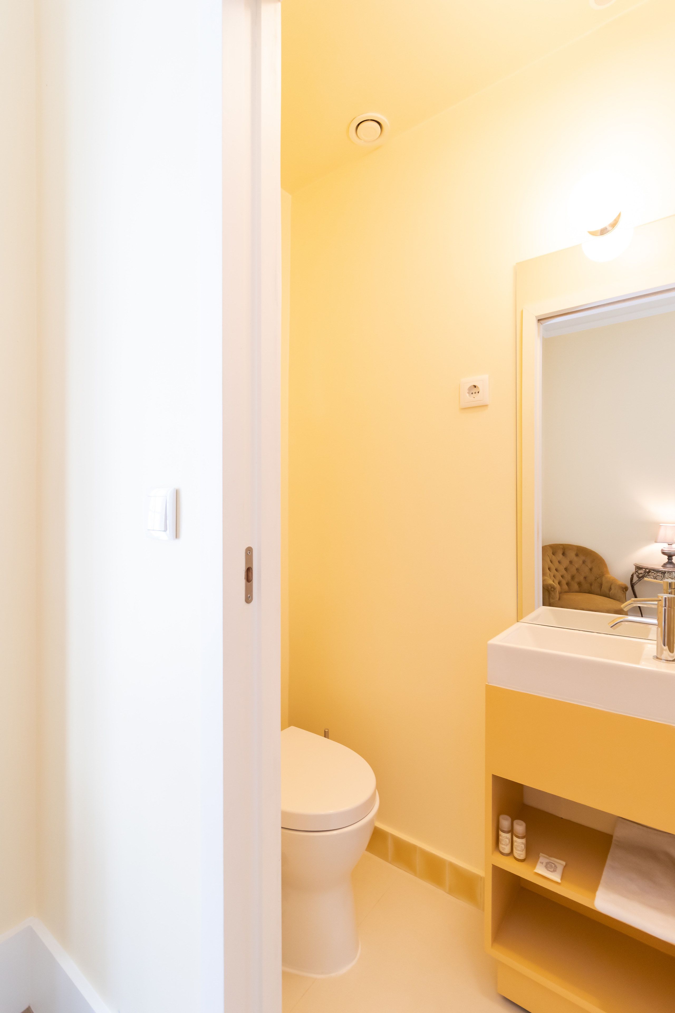 A bathroom with a white toilet, a yellow vanity with a mirror, and a shelf with toiletries.