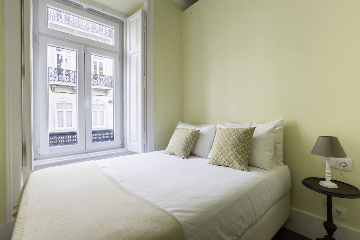 A bedroom with a bed, pillows, a bedside table with a lamp, and a window with a view of buildings.