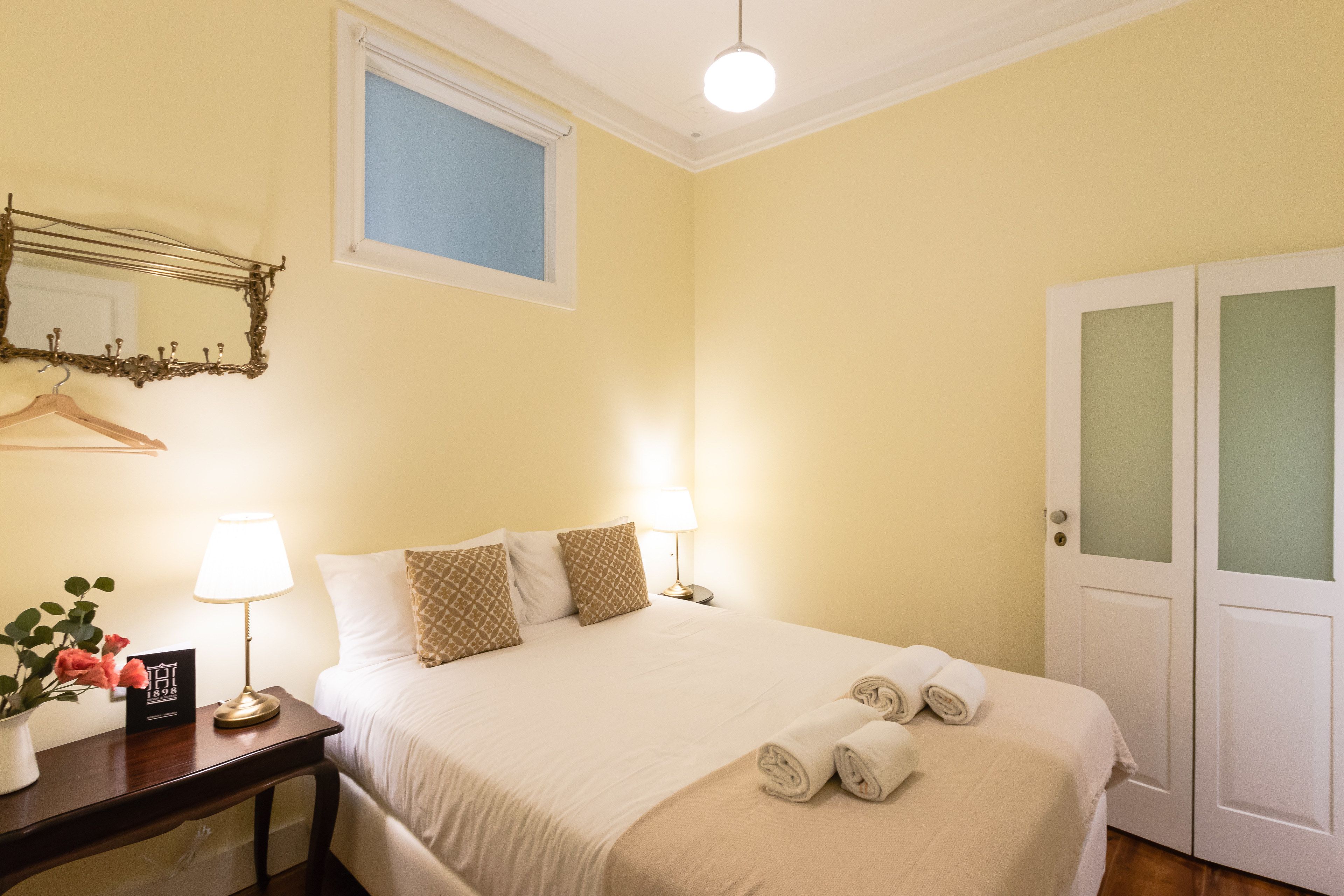 A neatly made bed with a white comforter and two pillows, a bedside table with a lamp and a small card, a mirror with a decorative frame, and a door with frosted glass panels.