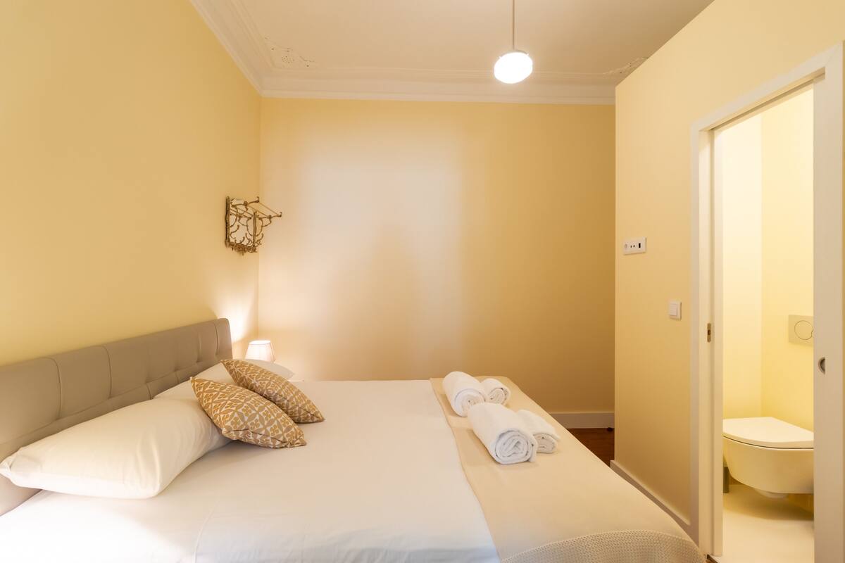 A neatly made bed with pillows and rolled towels, a bedside lamp, and a door leading to a bathroom.