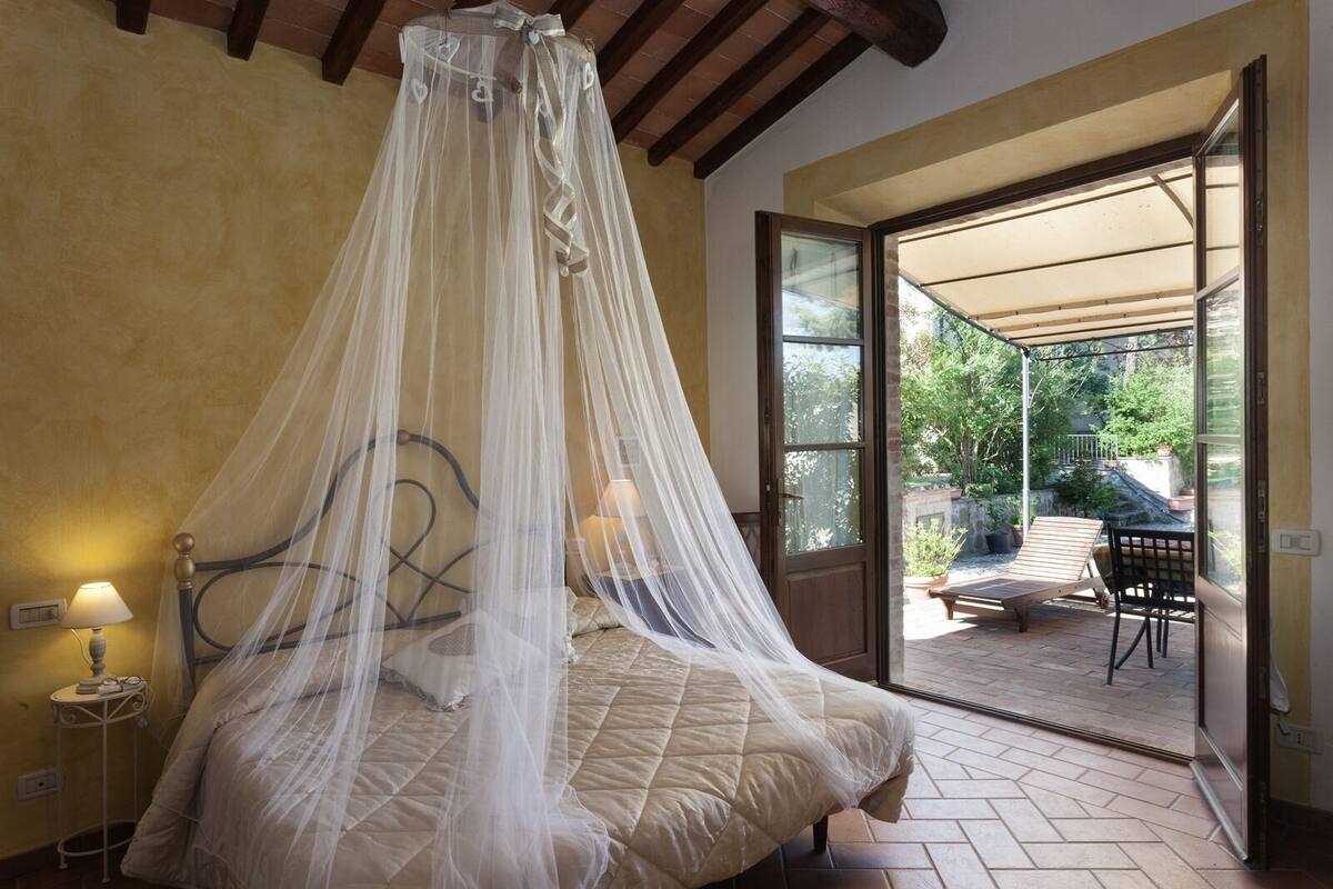 A bedroom with a canopy bed, a bedside table with a lamp, and a view of an outdoor patio with a table and chairs.