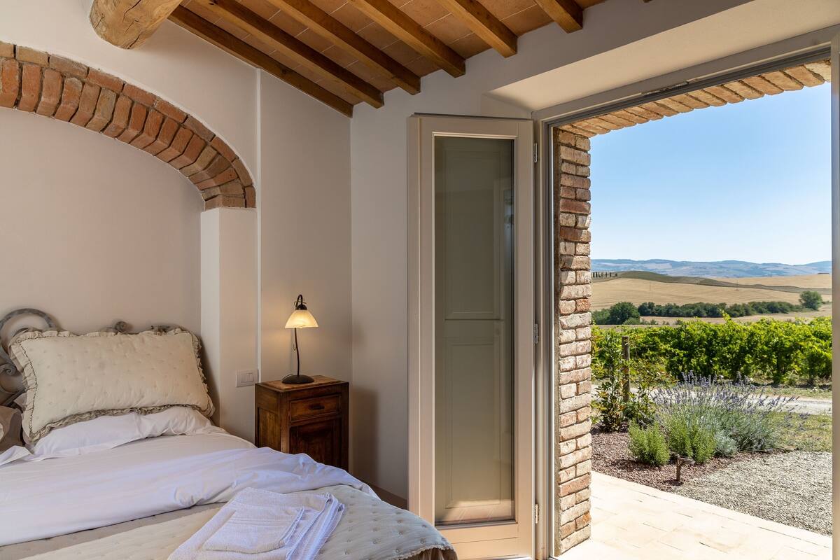 A bedroom with a bed, a nightstand, a lamp, and a view of a vineyard through an open door.