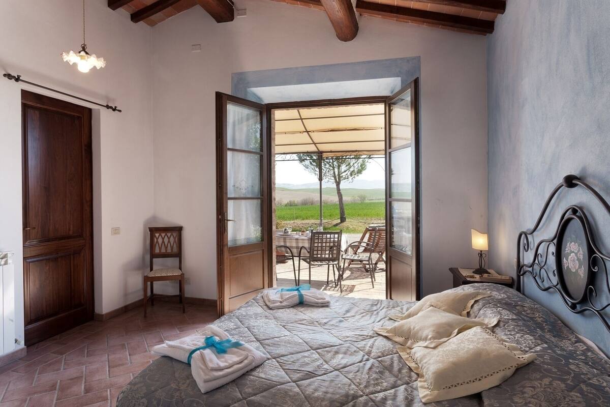 A bedroom with a bed, a wooden door, a chair, and an open door leading to an outdoor area with a table and chairs.