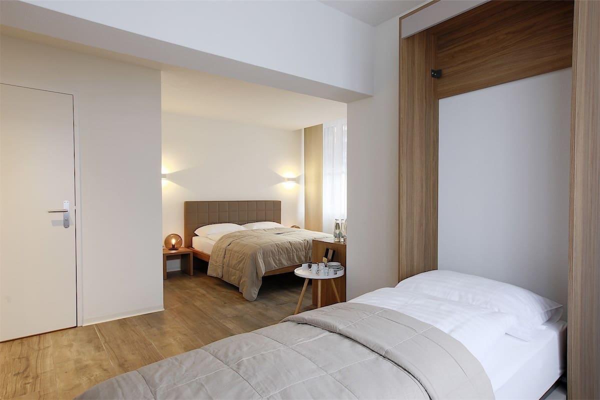 Una habitación de hotel moderna con una cama grande, una cama más pequeña, una mesita de noche con lámpara y un armario.