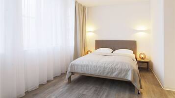 Una cama bien hecha con tablero de madera, dos lámparas de noche y cortinas traslúcidas sobre un piso de madera.