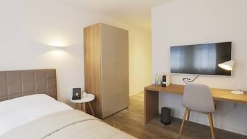 A modern hotel room with a bed, a desk with a computer, a TV, and a small side table with a lamp.