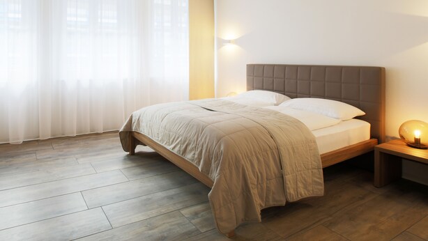 A bedroom with a bed, a nightstand with a lamp, and a window with sheer curtains. - Work Life Residence  (Winterthur)